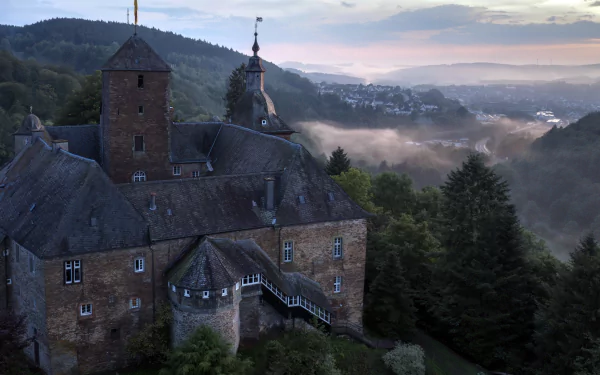 4K Ultra HD desktop wallpaper featuring a historic man-made castle perched on a hillside overlooking a misty forested valley at dawn.