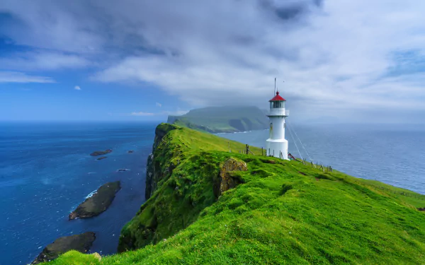 A vibrant 4K Ultra HD wallpaper of a white lighthouse on a lush green cliff overlooking the ocean in the Faroe Islands under a partly cloudy sky.