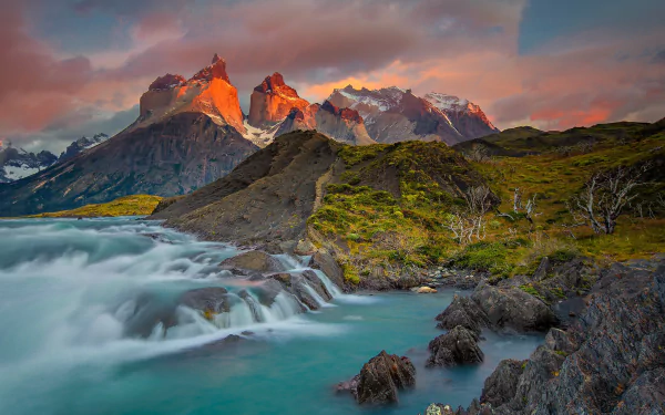 HD PC desktop wallpaper: Torres del Paine mountain peaks at sunset, dramatic cloud-lit summits above lush green hills, turquoise river with cascades in the foreground.