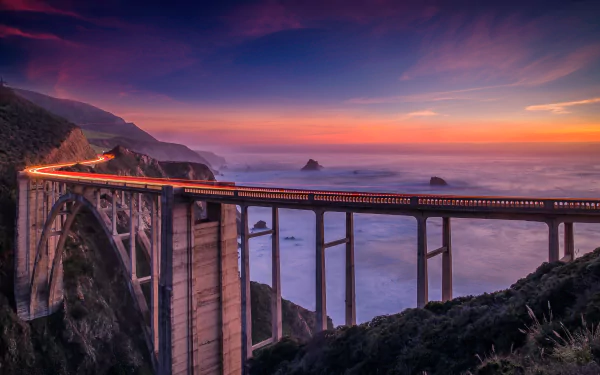 HD wallpaper of Bixby Creek Bridge on California's coast at sunset, with mountains, ocean fog, and a vibrant sky creating a picturesque landscape.