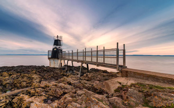 5K Ultra HD PC desktop wallpaper: man-made lighthouse on a rocky coast reached by a metal pier, pastel sunrise sky and smooth sea.