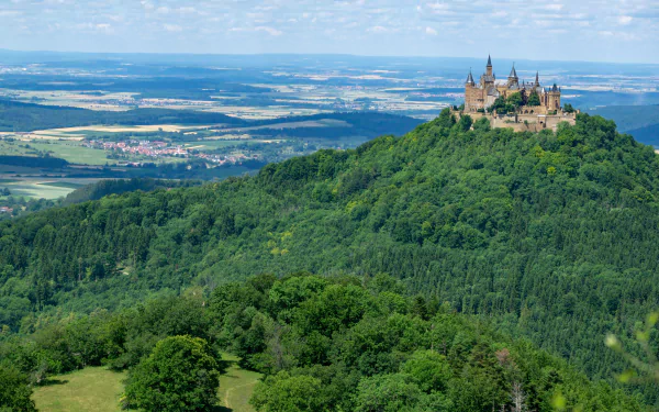 A panoramic 4K Ultra HD view of Hohenzollern Castle atop a lush forested hill in Germany, showcasing a scenic landscape with expansive fields and distant horizons.
