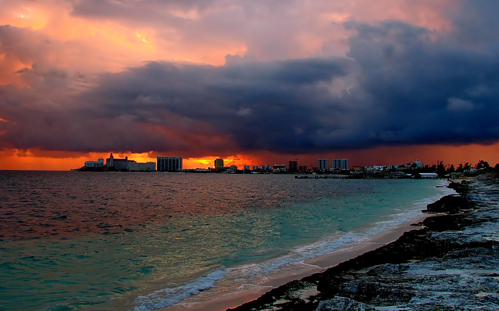 HD PC desktop wallpaper featuring a dramatic ocean shoreline at sunset with dark clouds and a city skyline in the distance, showcasing natural beauty and coastal vibes.