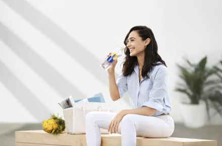 HD desktop wallpaper of smiling brunette Israeli actress and celebrity Gal Gadot sitting outdoors, holding a drink with a bright, minimal background and soft shadows.