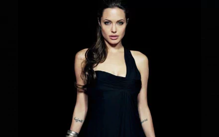 HD desktop wallpaper featuring celebrity Angelina Jolie. She is dressed in a black halter dress against a dark background, showcasing her tattoos and wearing silver bracelets.