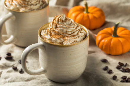 HD desktop wallpaper of a cozy autumn scene with hot coffee topped with whipped cream, surrounded by coffee beans and small pumpkins.