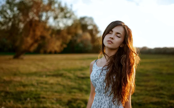 HD desktop wallpaper showing a brunette woman outdoors, exuding a serene mood with the sun setting in the background and a green, grassy field.