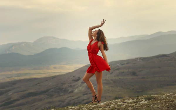 HD desktop wallpaper of a woman in a red dress striking a graceful profile pose atop a hill, with muted mountains and moody skies in the background.