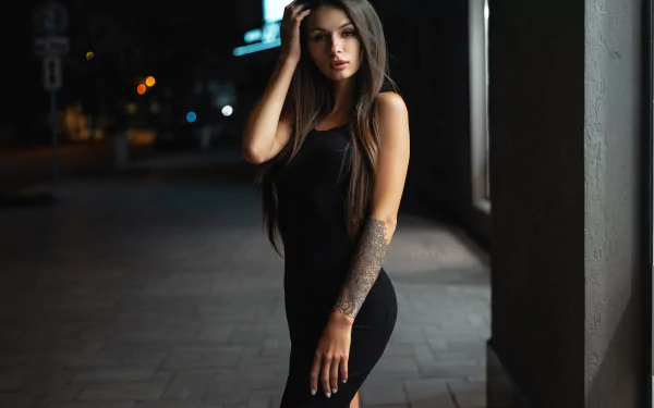 Long-haired woman/model in a black dress with a forearm tattoo, posed against a dim urban night backdrop — 2K Quad HD PC desktop wallpaper.
