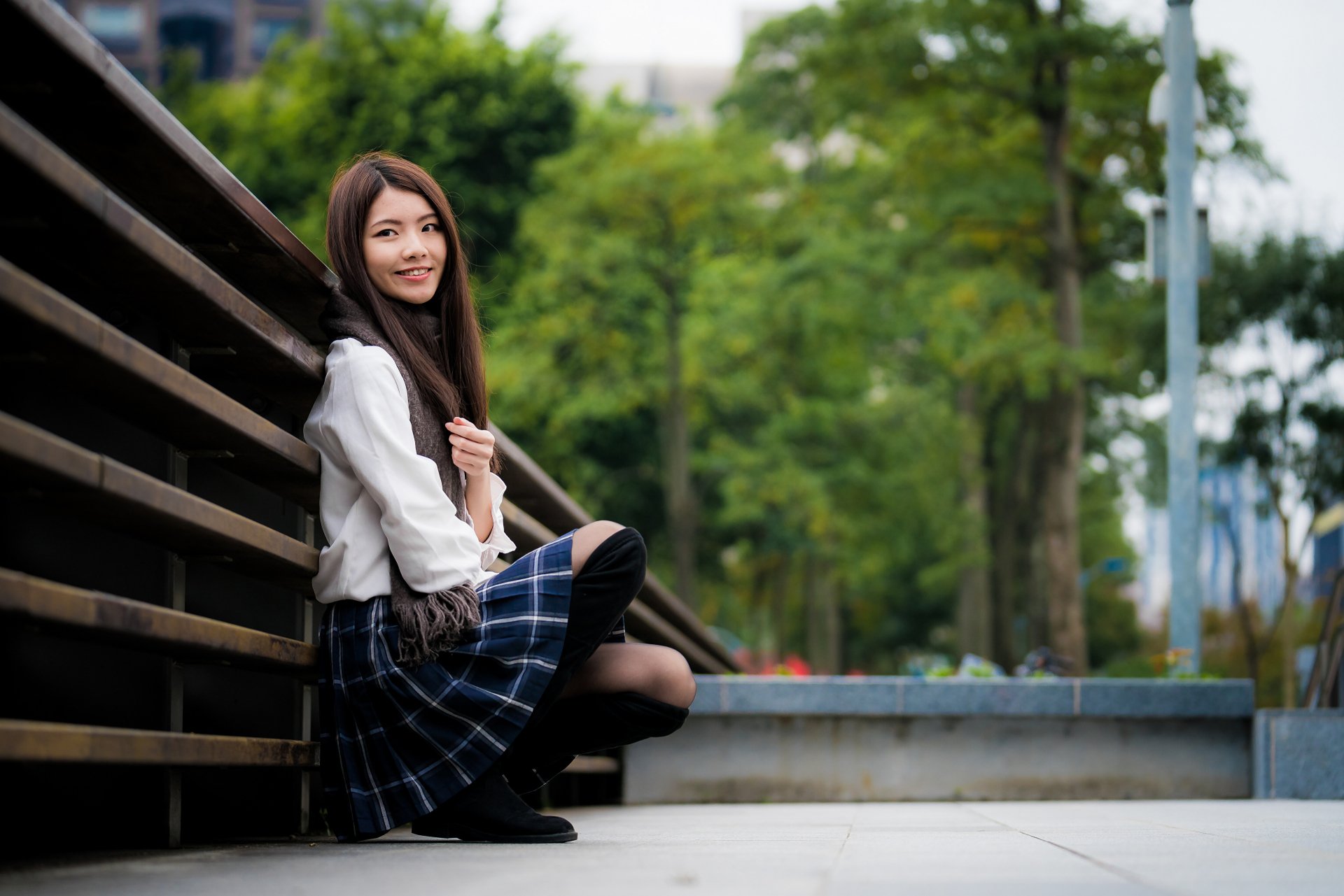A brunette Asian woman model with a plaid skirt and white top sits outdoors, captured with a shallow depth of field in this 4K Ultra HD desktop wallpaper.