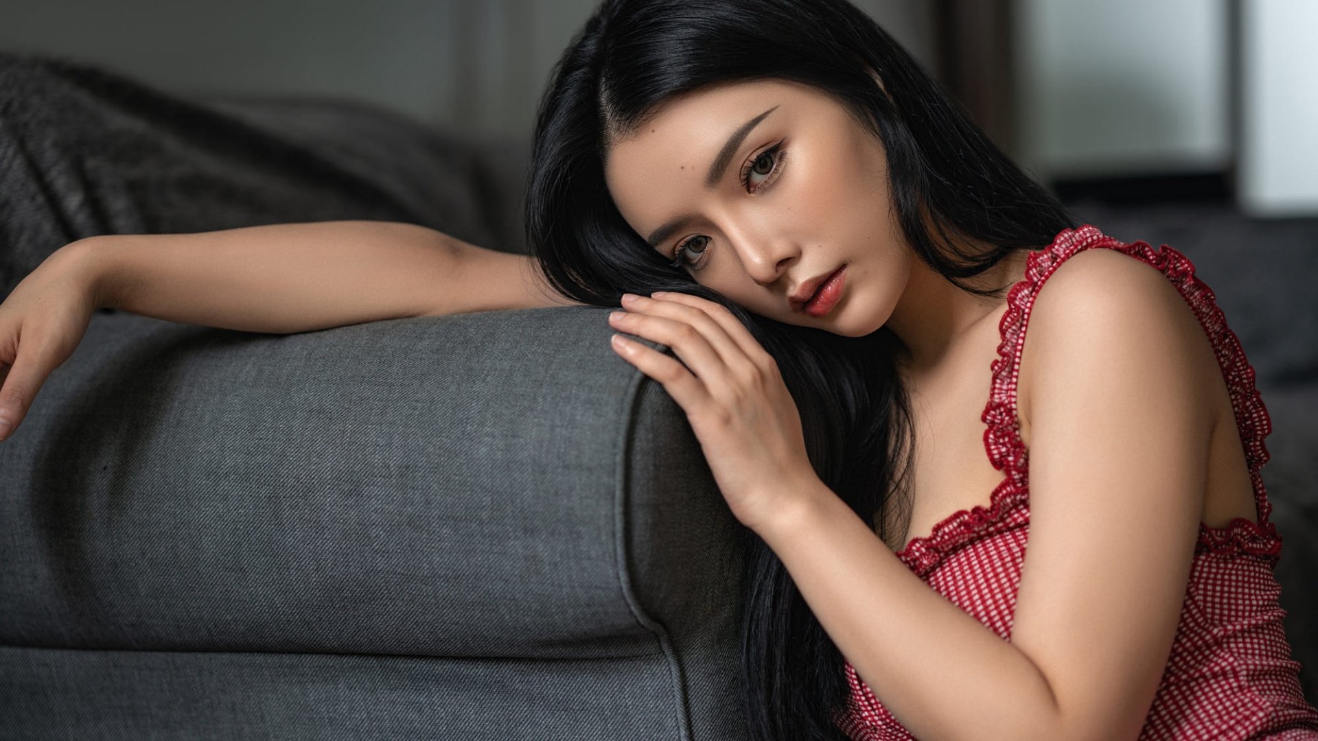 HD desktop wallpaper featuring an Asian woman model with long black hair, resting her head on a gray sofa, wearing a red patterned top.