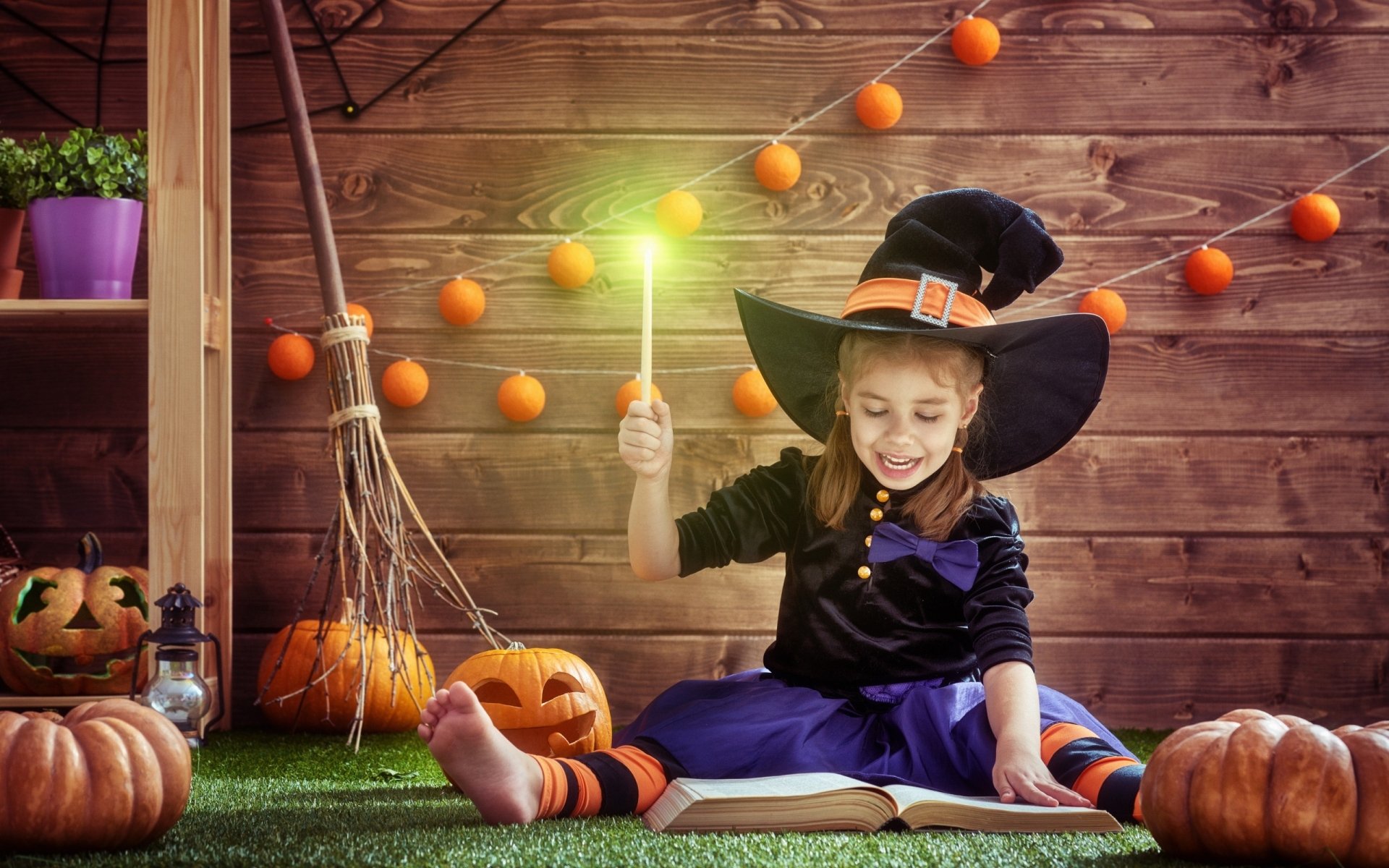 A child dressed as a witch with bare feet, holding a glowing wand, surrounded by pumpkins and Halloween decorations on a wooden floor, captured in vibrant HD photography.