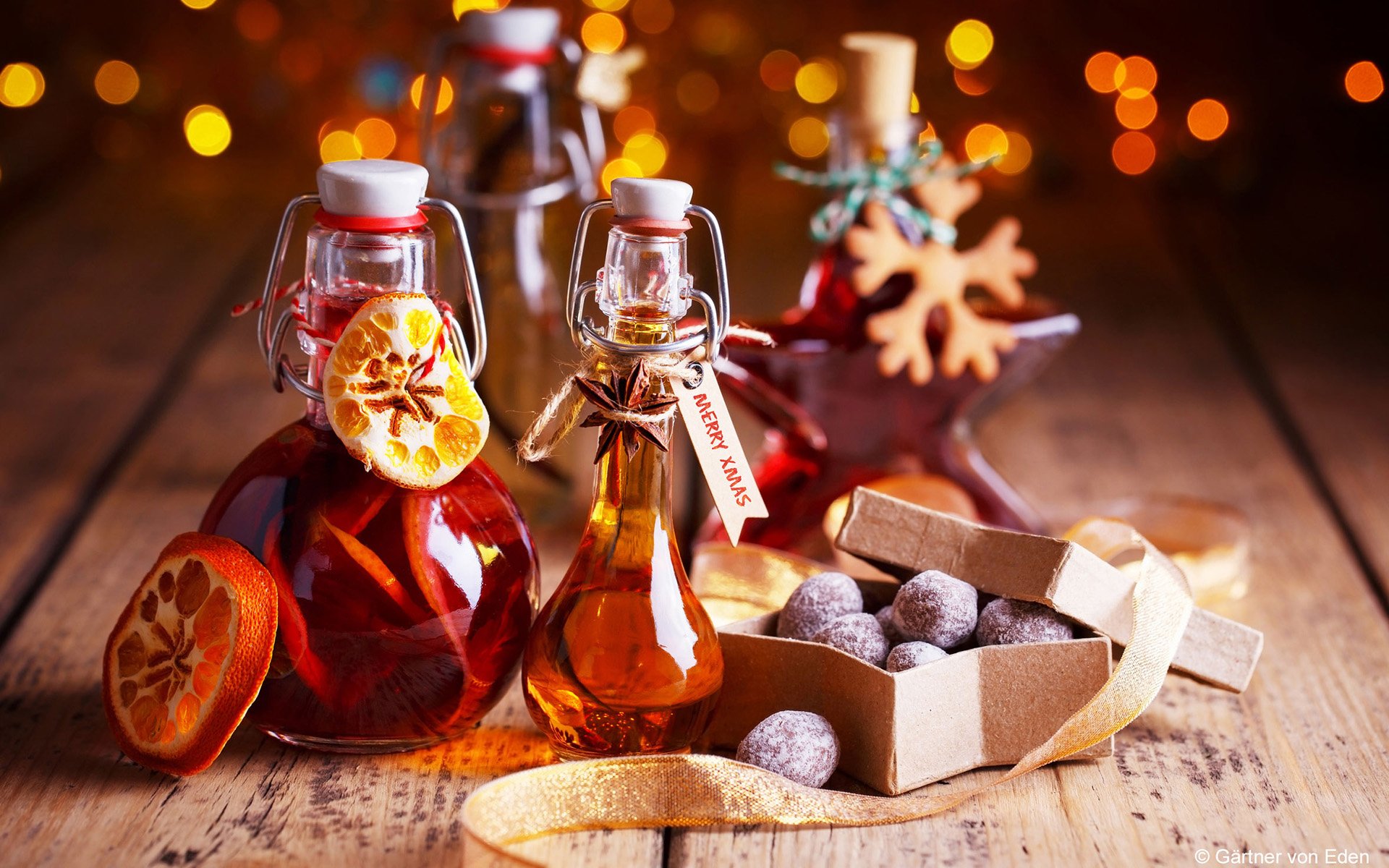 HD desktop wallpaper of Christmas mulled wine in glass bottles with dried citrus, candy and chocolate truffles on a wooden table, warm bokeh lights in the background.