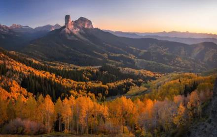 HD PC desktop wallpaper: autumn forest with golden aspens in foreground, pine-clad valleys and a rugged mountain beneath a pastel sunrise sky.