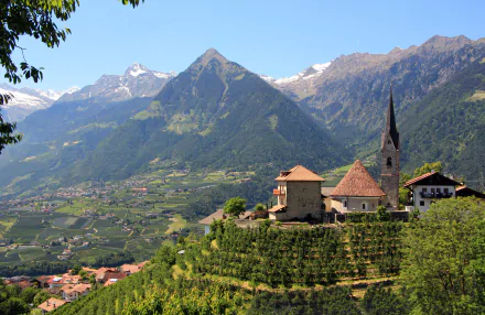 Panoramic HD desktop wallpaper of a valley in South Tyrol, Italy, featuring a mountain backdrop and a historic church nestled among vineyards.