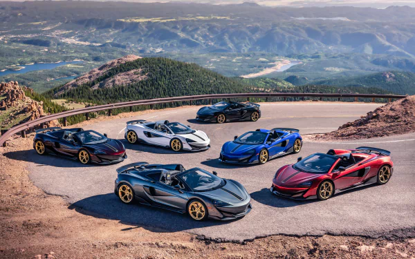 A lineup of six McLaren 600LT supercars in various colors, parked on a mountain road with a scenic mountain landscape in the background, rendered in 4K Ultra HD.