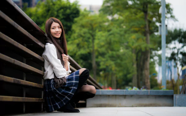 A brunette Asian woman model with a plaid skirt and white top sits outdoors, captured with a shallow depth of field in this 4K Ultra HD desktop wallpaper.