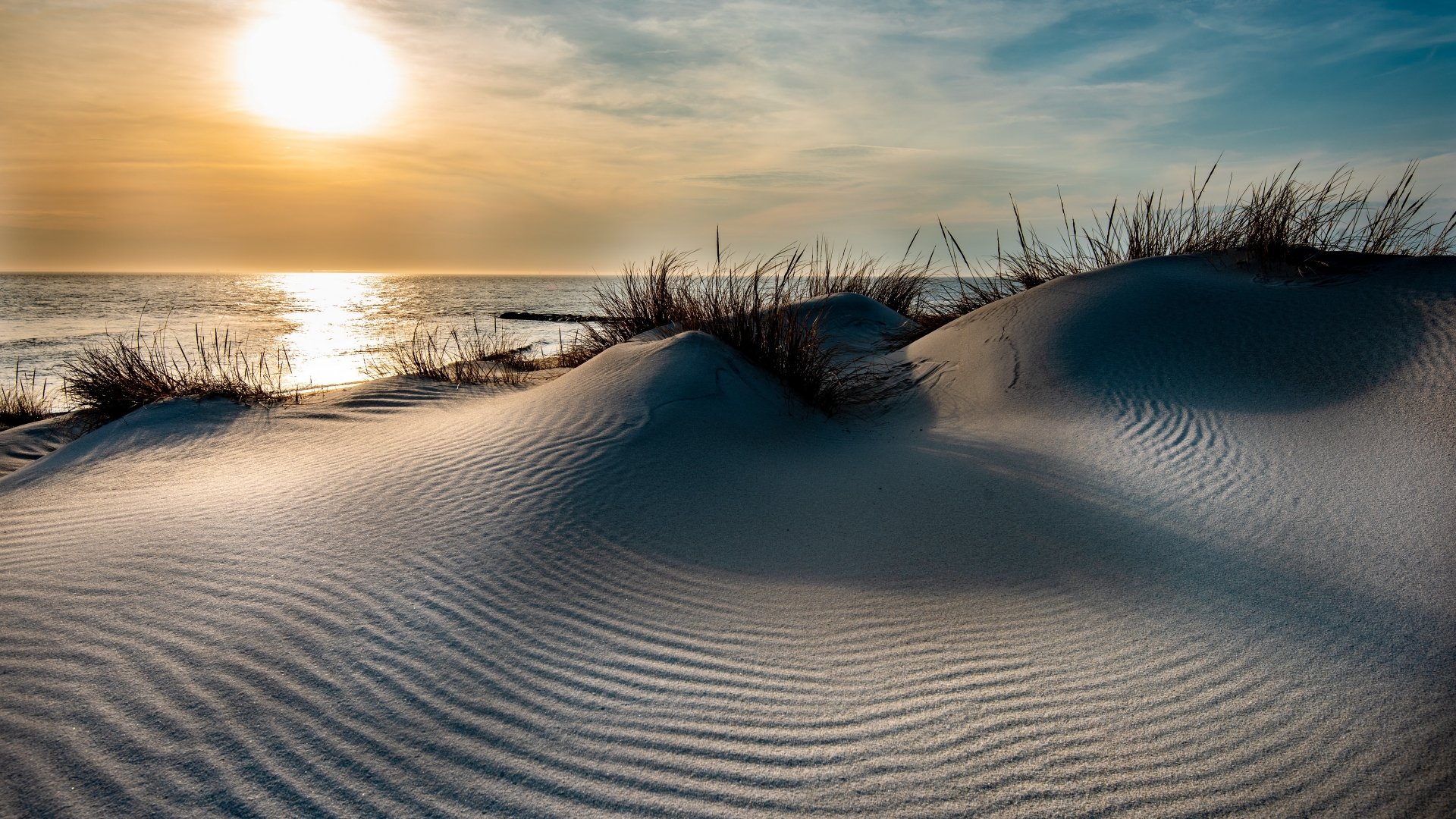 4K Ultra HD PC desktop wallpaper: rippled sand dunes and beach grass at sunset overlooking the sea, golden sun reflecting on water and sky, a serene nature scene.