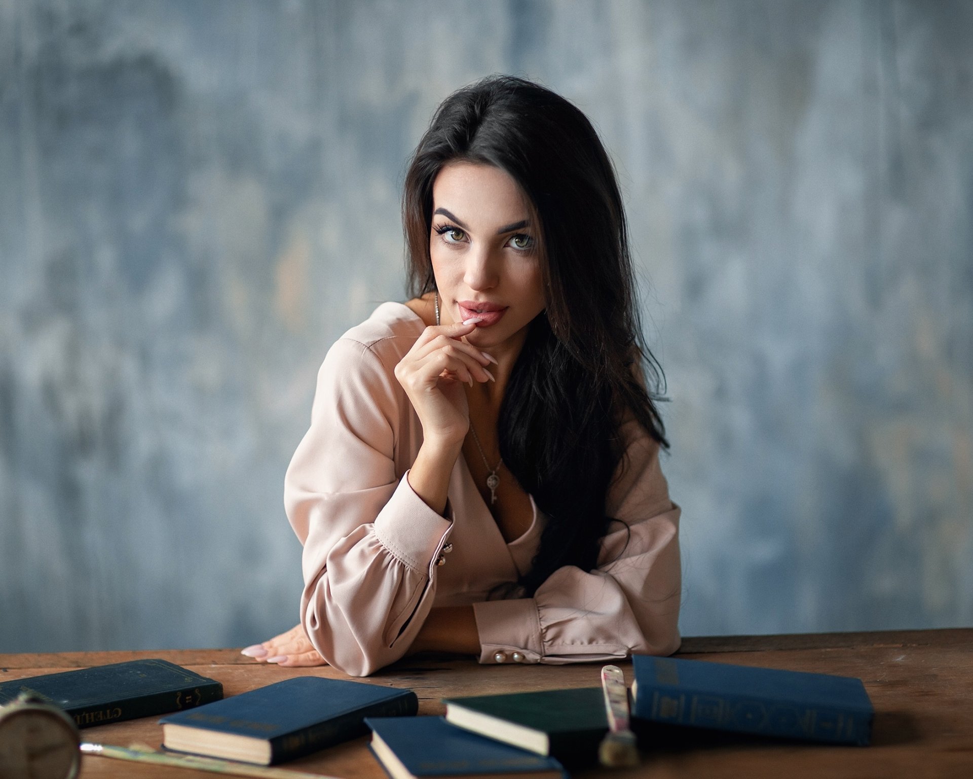HD PC desktop wallpaper of a black‑haired woman model with long hair, leaning on a table and gazing at the camera against a soft gray textured background.