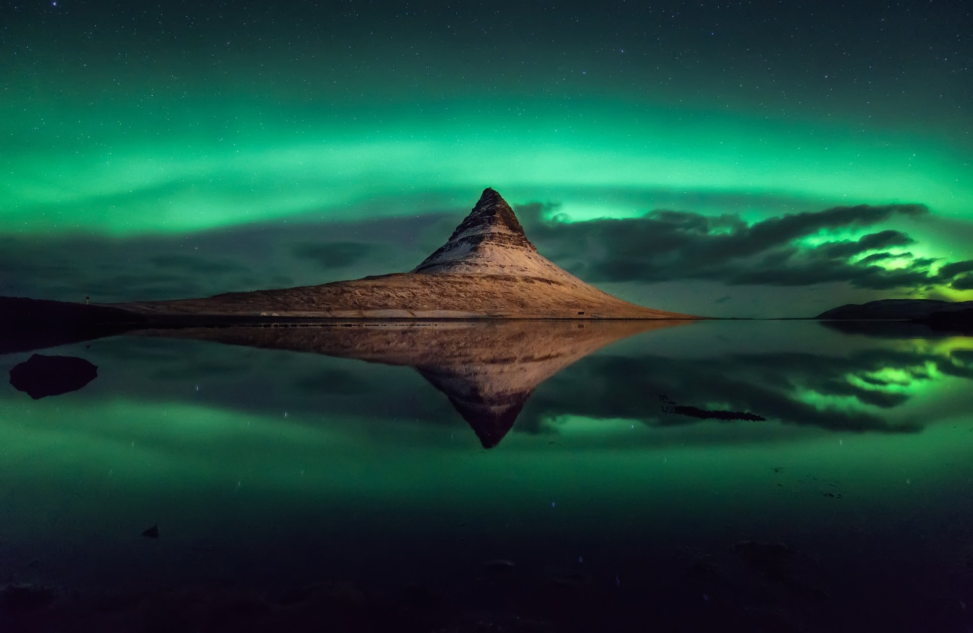 Aurora borealis lights up the night sky over Kirkjufell mountain in Iceland, beautifully reflected in the calm water under a starry sky.