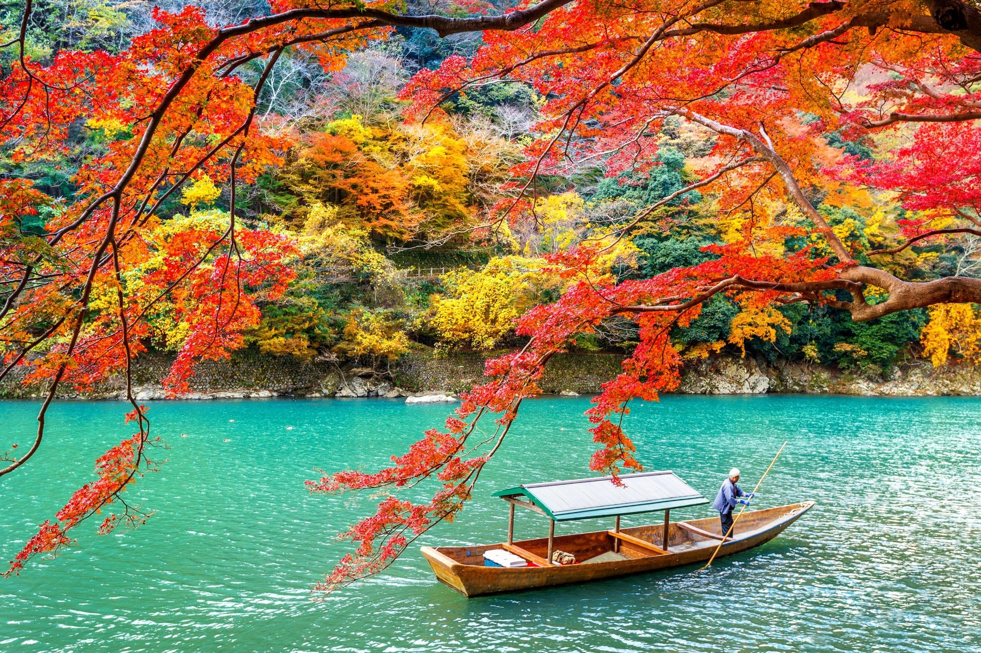 A serene 4K Ultra HD view of Arashiyama park’s vibrant fall foliage over a turquoise lake in Kyoto, Japan, with a boat gently floating in the water.
