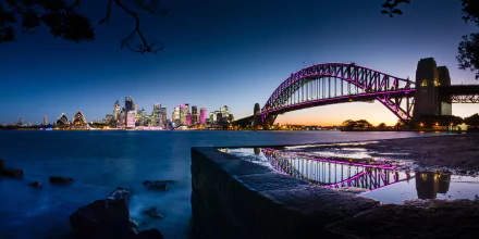 5K UHD PC wallpaper of Sydney Harbour Bridge at dusk, purple-lit arch and city skyline reflecting on water in New South Wales, Australia — a man-made bridge and shoreline.