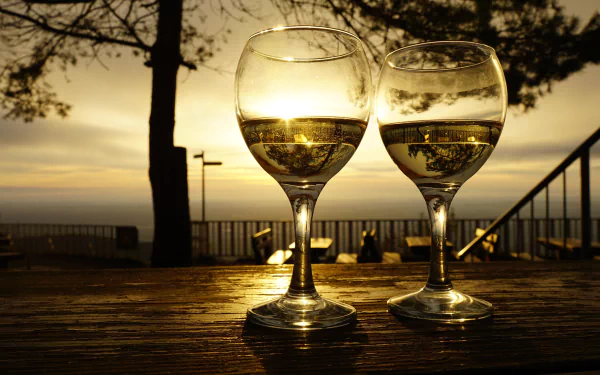 Two wine glasses filled with white wine on a wooden table at sunset, overlooking a scenic outdoor view; 4K Ultra HD PC desktop wallpaper capturing a warm, golden ambiance.