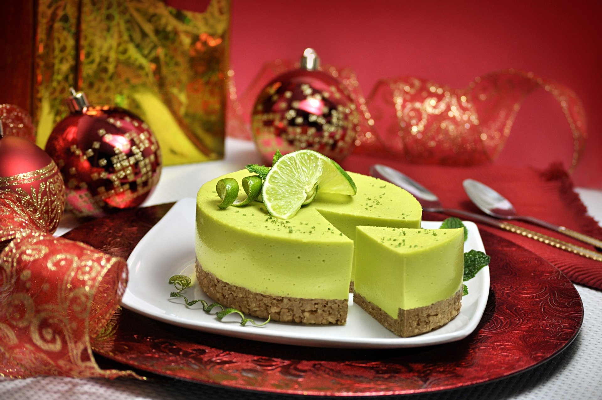 HD desktop wallpaper of a vibrant lime cheesecake with a crumb crust, garnished with lime slices, set against a festive red background with ornaments and ribbon.