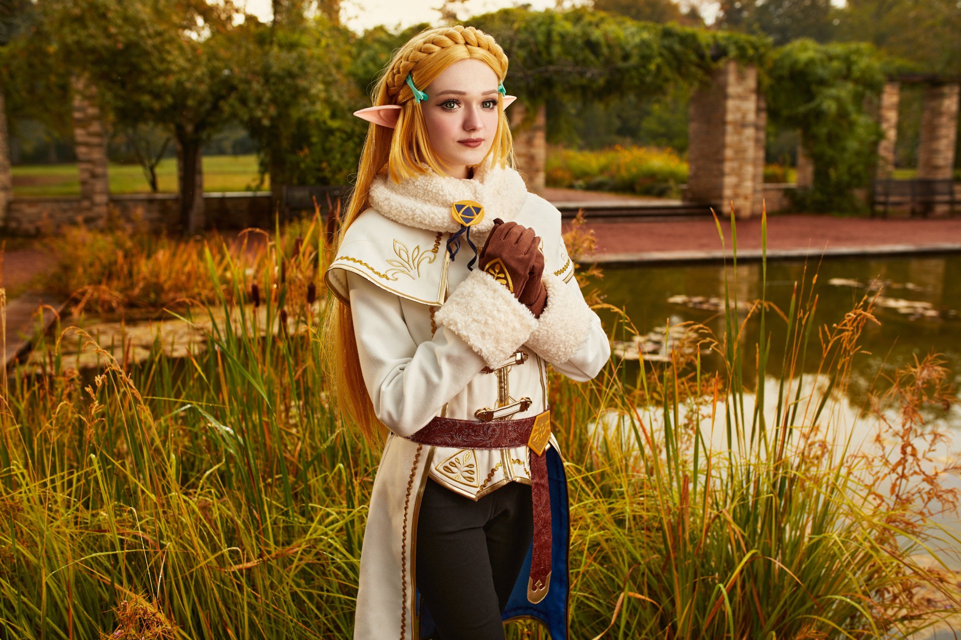 Cosplay of Princess Zelda from The Legend of Zelda, featuring a blonde elf woman in detailed costume standing by a pond with autumn foliage, captured in 4K Ultra HD.