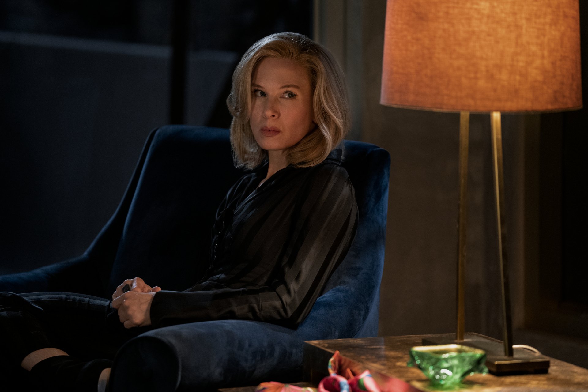 HD desktop wallpaper featuring Renée Zellweger from the series What/If, sitting pensively in a dark room lit by a lamp.