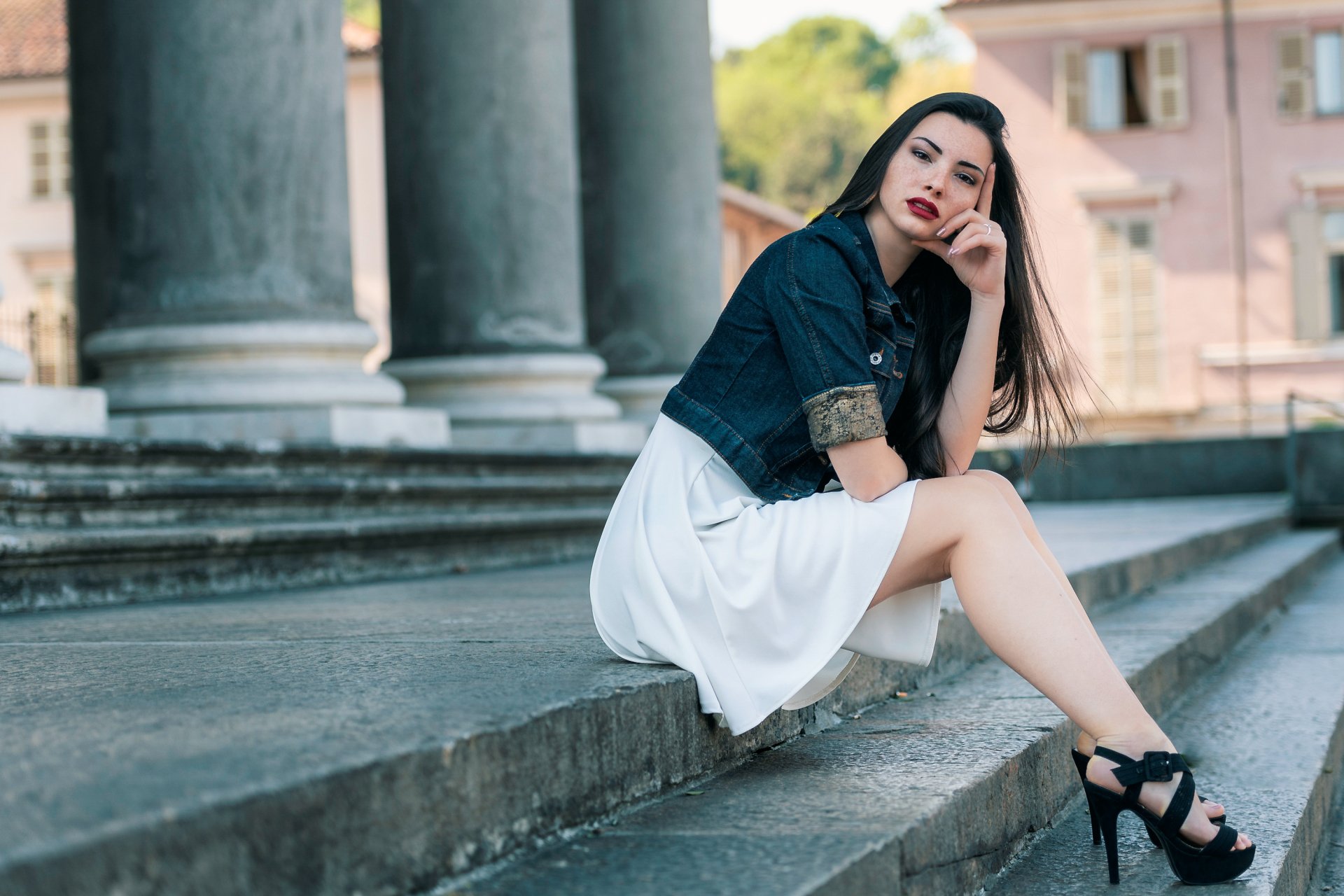 Depth-of-field portrait of a black-haired woman model in a denim jacket and white dress, red lipstick, seated on stone steps — 2K Quad HD PC desktop wallpaper background.