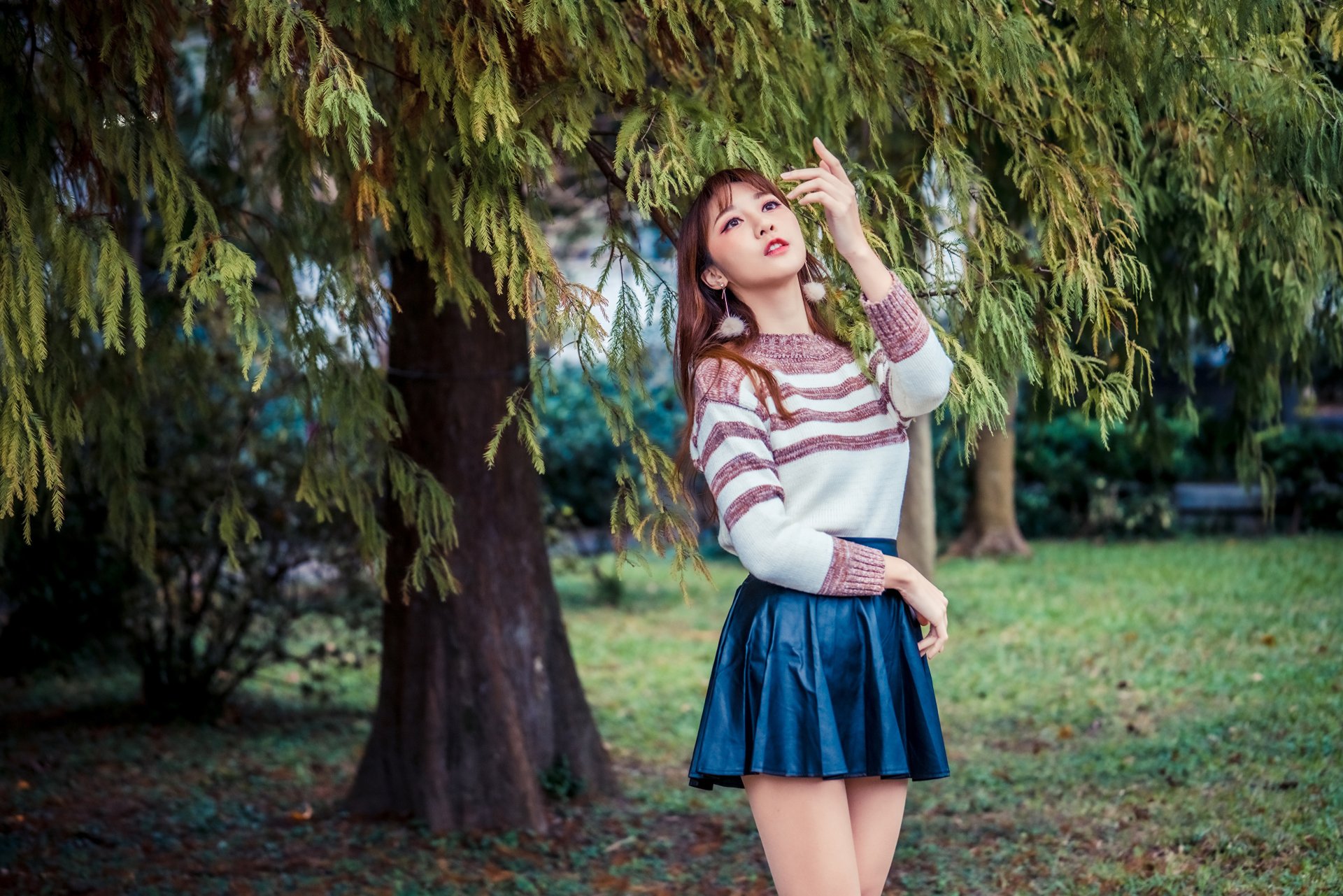 Brunette Asian woman in a striped sweater and navy skirt poses under lush green trees in a serene outdoor setting, captured in 4K Ultra HD for desktop wallpaper.
