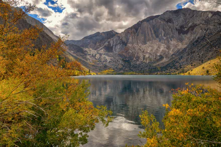 8K Ultra HD image of a tranquil mountain lake surrounded by autumn foliage under dramatic clouds, showcasing pristine water and stunning natural outdoor scenery.