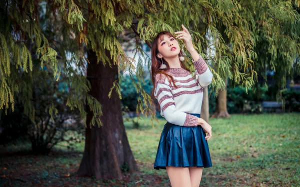 Brunette Asian woman in a striped sweater and navy skirt poses under lush green trees in a serene outdoor setting, captured in 4K Ultra HD for desktop wallpaper.
