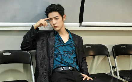 Seated young man in a teal patterned shirt and black blazer, hand to temple, posed against folding chairs — 2K Quad HD PC desktop wallpaper/background.