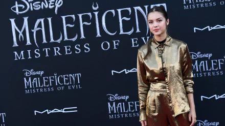 Brunette American singer and actress Olivia Rodrigo poses at the Maleficent: Mistress of Evil premiere, dressed in a shiny bronze outfit against a branded backdrop.