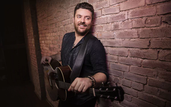 Smiling guitarist leaning against a brick wall holding a guitar, suitable as HD desktop wallpaper and background.