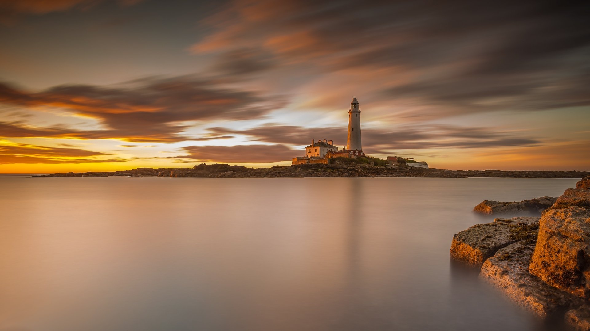 A 4K Ultra HD desktop wallpaper featuring a man-made lighthouse on a small island under a dramatic sunset sky with smooth water and rocky foreground.