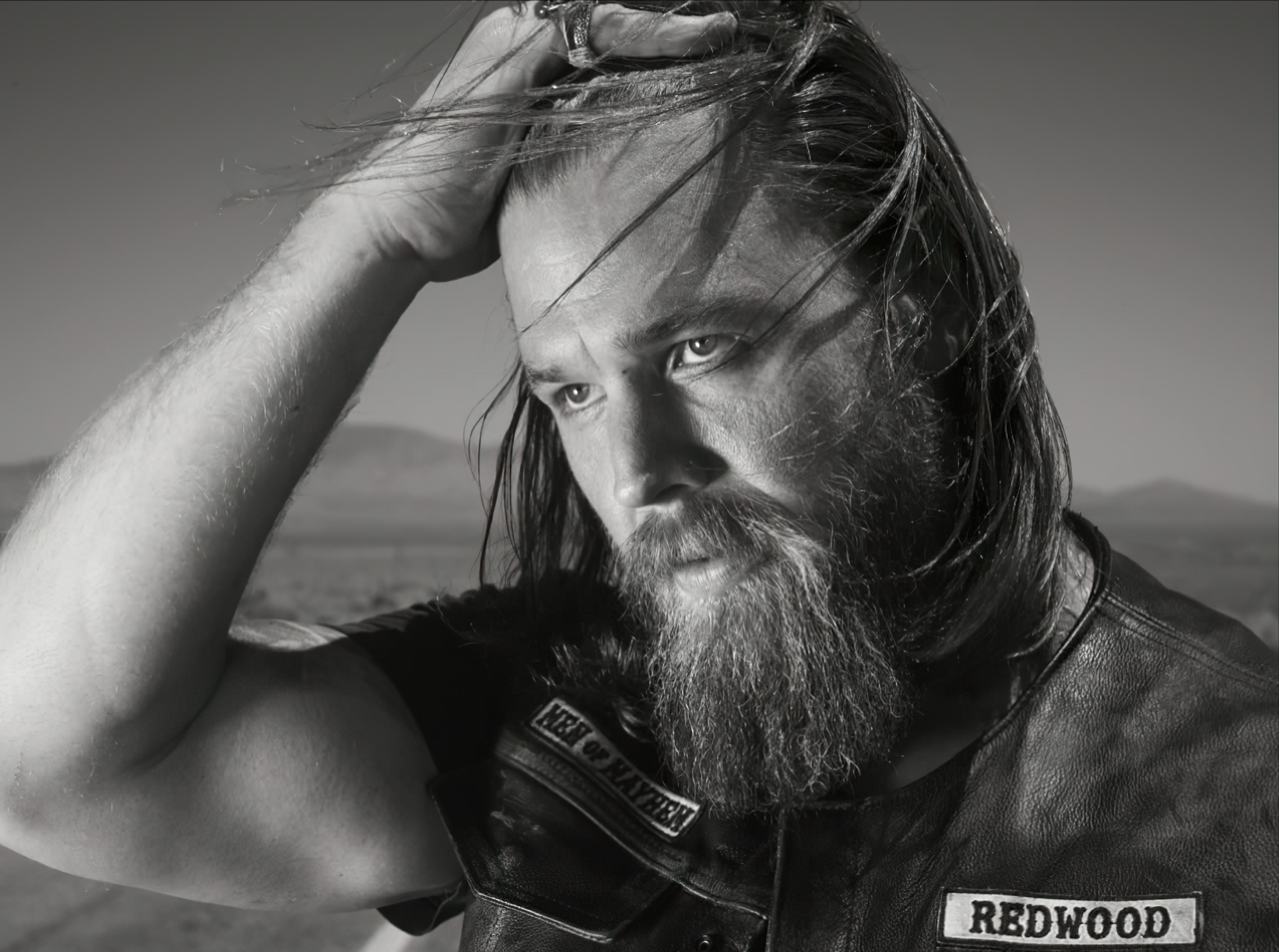 Black and white HD wallpaper featuring a bearded man in a leather vest, reminiscent of a Sons of Anarchy character, posing against a blurred natural backdrop.