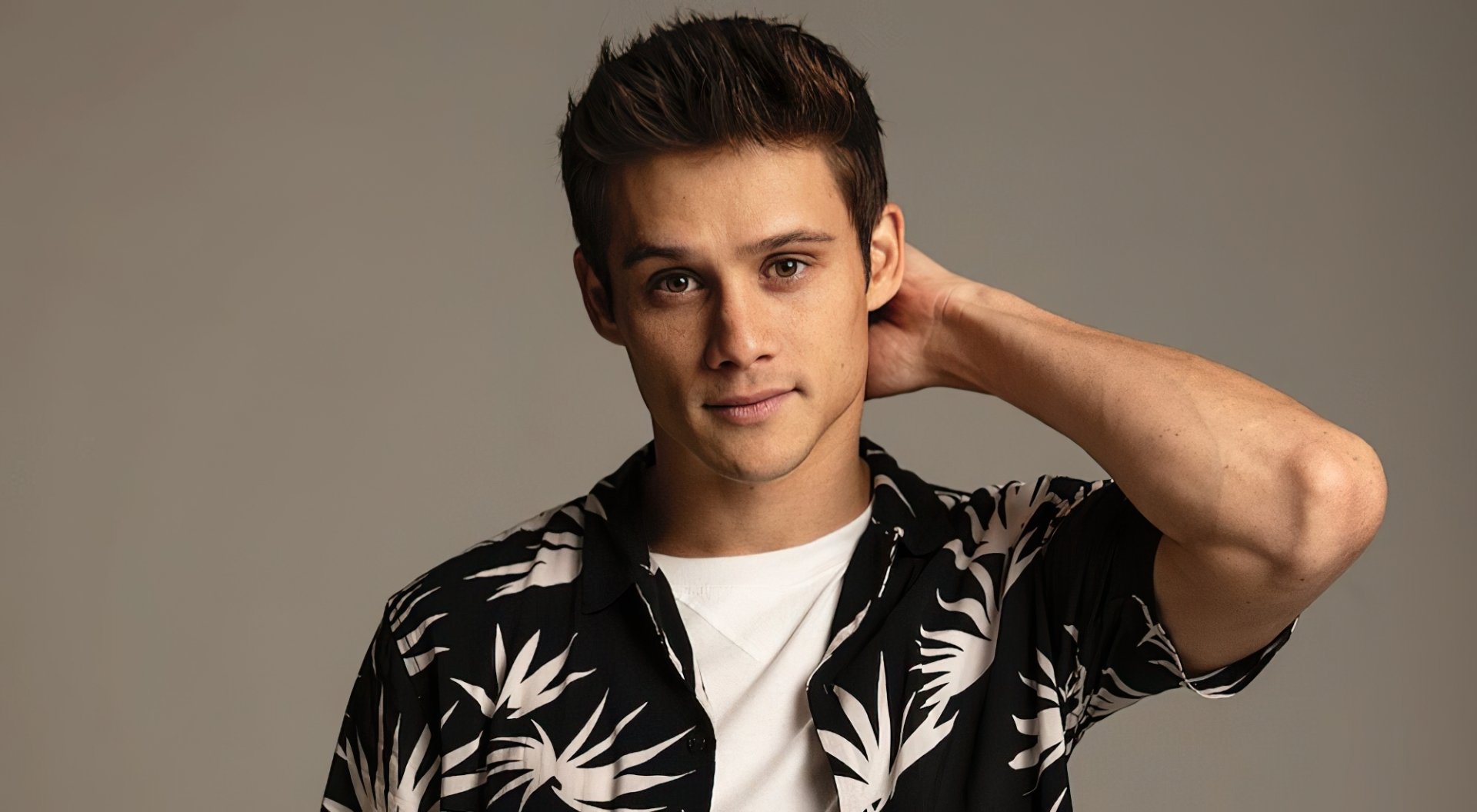 HD desktop wallpaper featuring a confident young man with a stylish haircut, wearing a black and white tropical print shirt, posing with his hand behind his head.