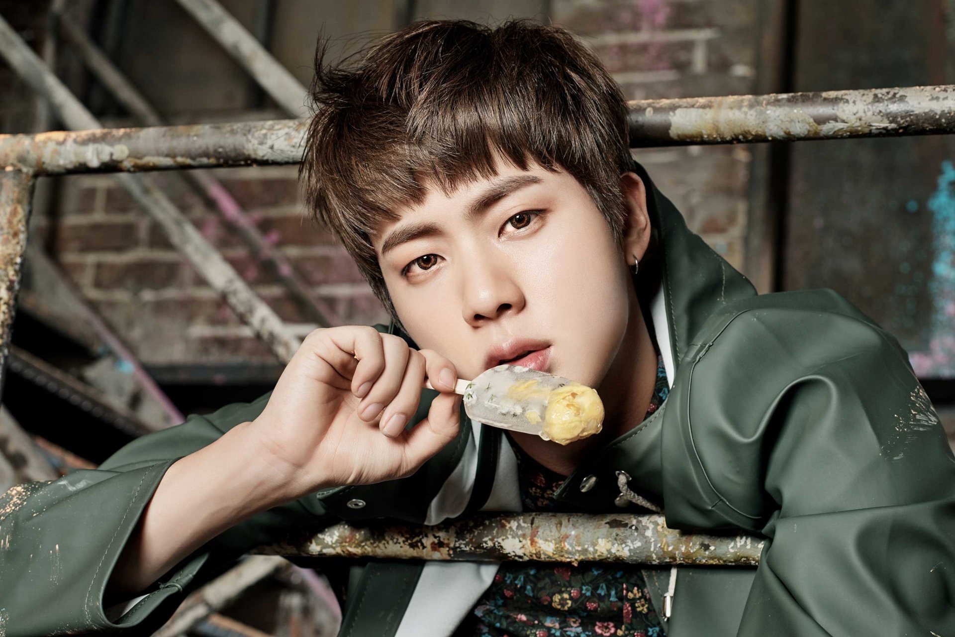 HD desktop wallpaper: close-up of a K-pop singer leaning on metal scaffolding, eating a popsicle, soft lighting and textured industrial background.