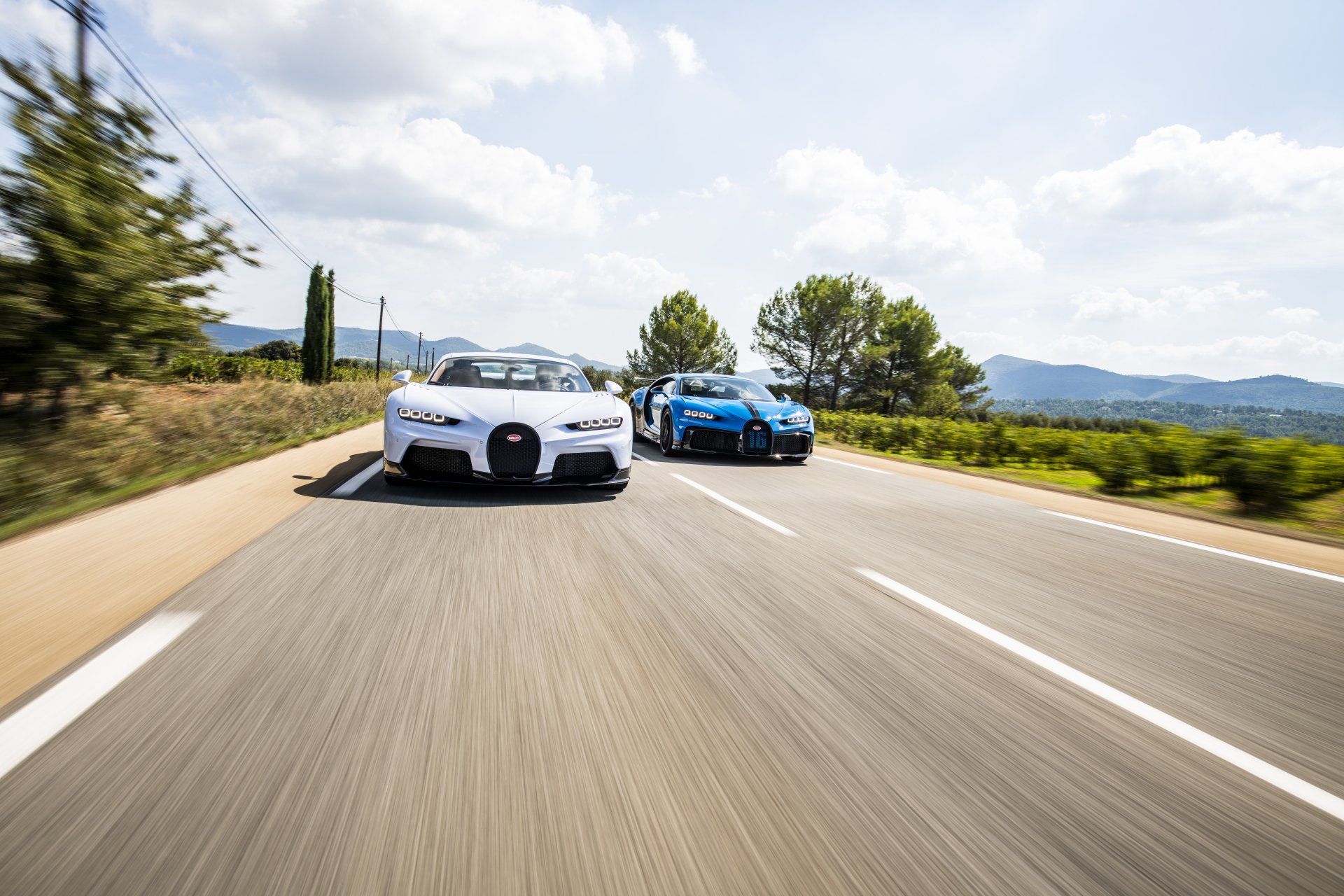 Two Bugatti Chiron supercars speeding down a sunlit open road — 8K Ultra HD PC desktop wallpaper background of high-performance vehicles.