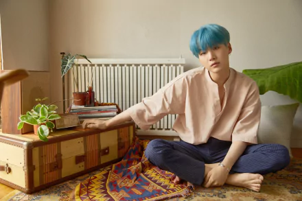4K Ultra HD wallpaper of Suga (Min Yoongi) from BTS, sitting barefoot on a colorful rug in a cozy room with plants and books, showcasing a calm and relaxed vibe.
