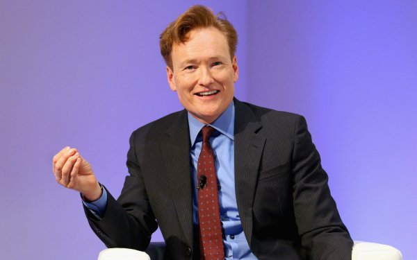 Smiling man with red hair wearing a suit and blue tie seated in front of a lavender background, ideal for HD desktop wallpaper and background.