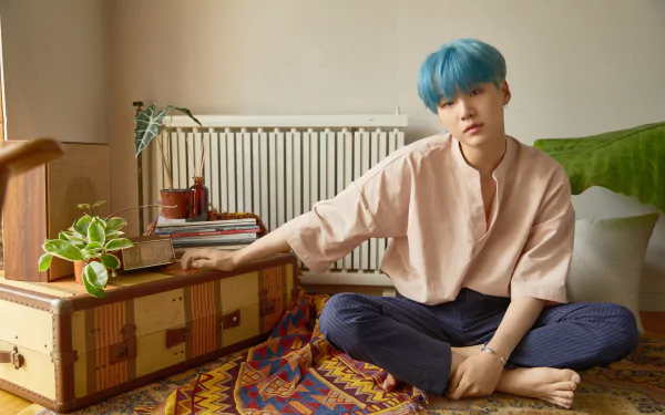 4K Ultra HD wallpaper of Suga (Min Yoongi) from BTS, sitting barefoot on a colorful rug in a cozy room with plants and books, showcasing a calm and relaxed vibe.
