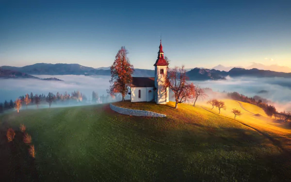 HD desktop wallpaper of a serene religious church atop a hill in Slovenia, surrounded by misty fields and a soft sunrise glow.