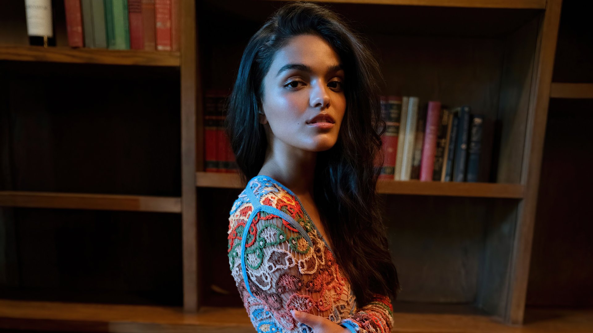 Portrait of celebrity Rachel Zegler with black hair, set against a dimly lit bookshelf background, captured in 4K Ultra HD for a PC desktop wallpaper.