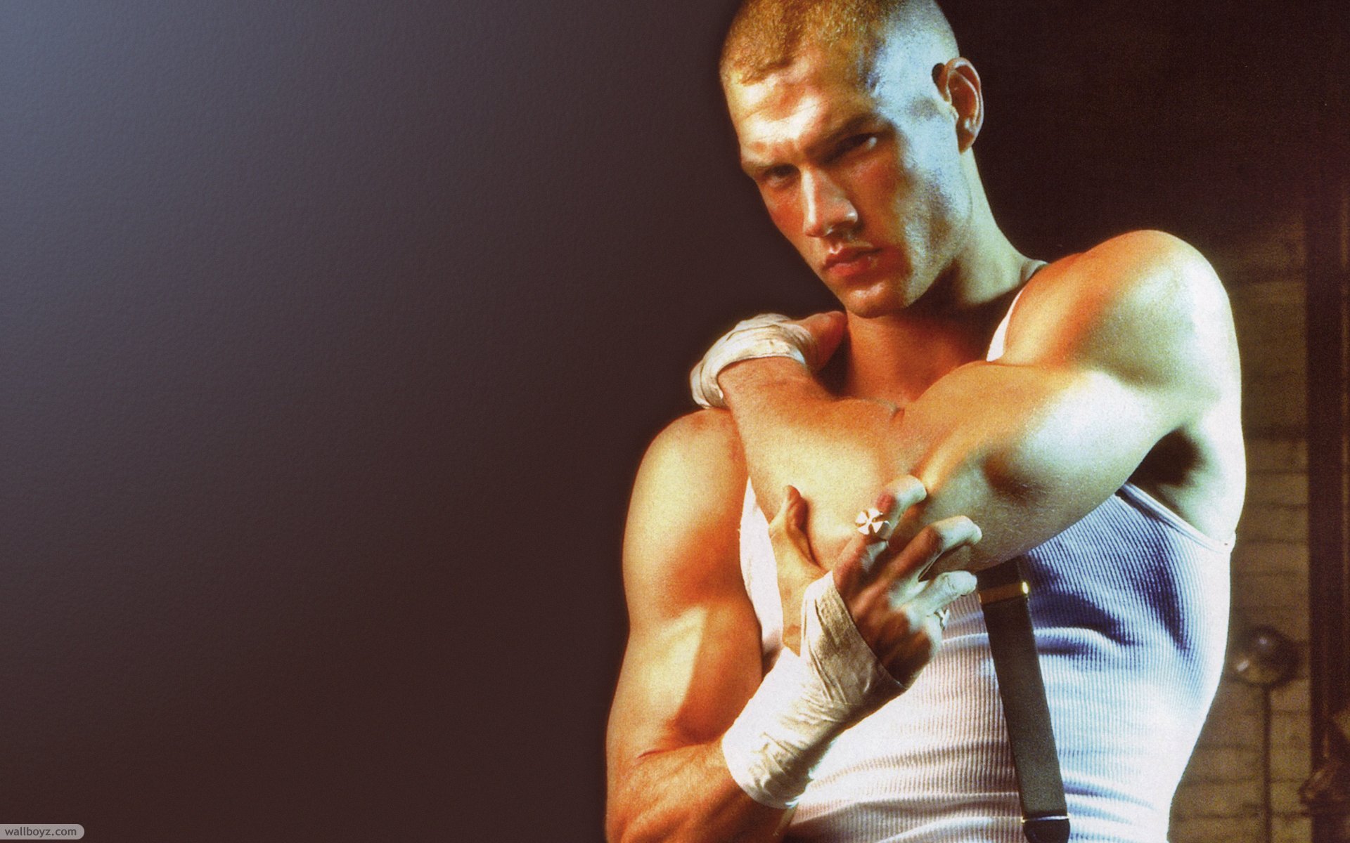 A muscular man in a tank top and suspenders poses with a serious expression, showcasing athleticism and confidence. This HD PC desktop wallpaper features a striking and dramatic background.