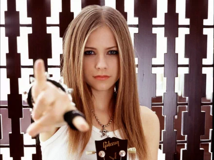 A young woman with long hair points forward confidently, set against a patterned background. This HD wallpaper features Avril Lavigne, embodying a bold music vibe.