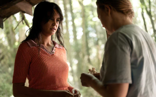 Two women converse beneath trees, one in an orange knit top and one in a gray tee; sunlit forest scene — Yellowjackets TV show 4K Ultra HD PC desktop wallpaper.